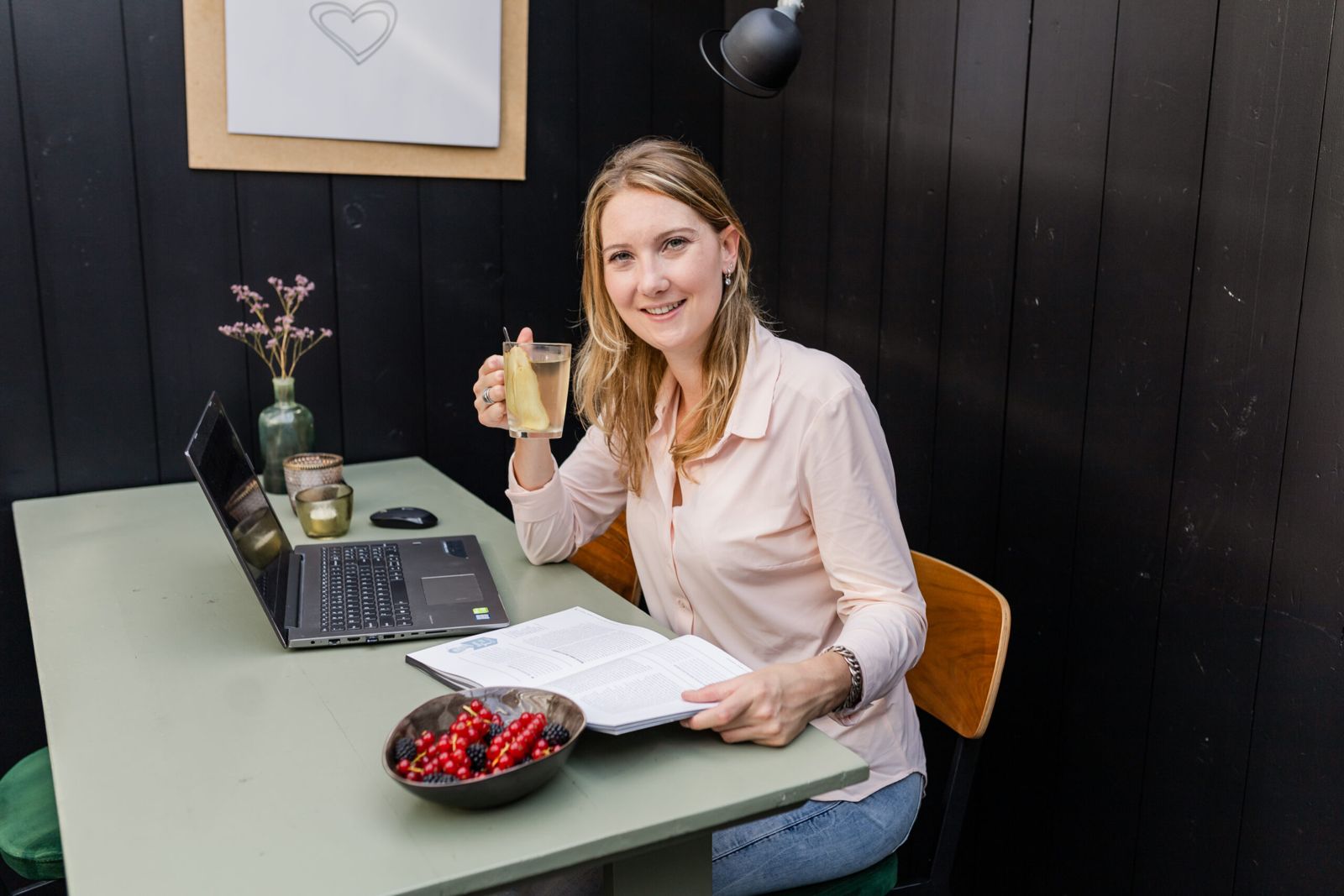 Wendy Reijnders met een kop thee aan haar bureau, met laptop, boek en een schaal bessen